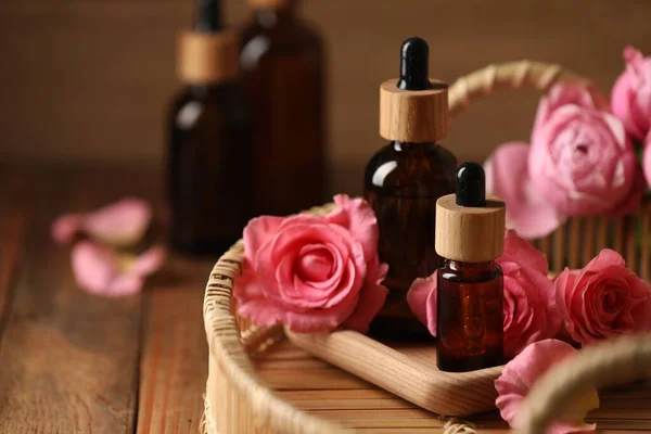 Tray with bottles of essential rose oil and flowers on wooden table, closeup