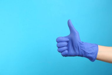 Tıbbi eldivenli biri açık mavi arka planda baş parmağını gösteriyor, eli yakın. Metin için boşluk