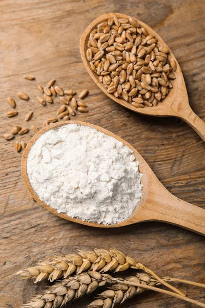 Spoons with wheat flour, grains and spikes on wooden table, above view