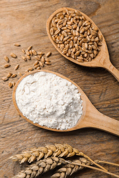 Spoons with wheat flour, grains and spikes on wooden table, above view