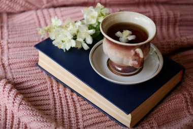Cup of aromatic tea with beautiful jasmine flowers and book on pink fabric