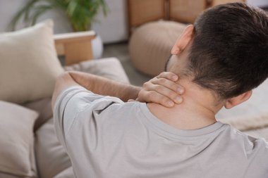 Man suffering from neck pain in living room, back view