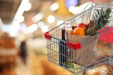Shopping cart with different groceries in supermarket, space for text