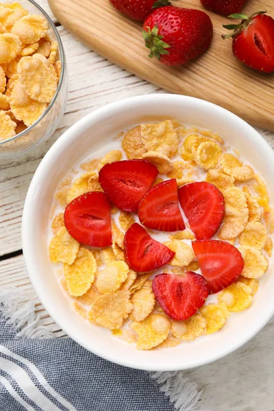 Bowl of tasty crispy corn flakes with milk and strawberries on white wooden table, flat lay
