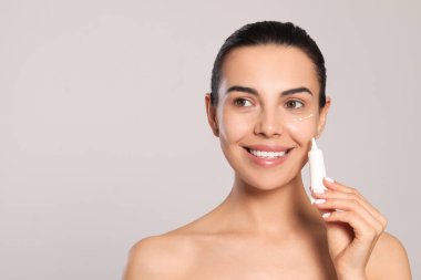 Beautiful young woman with cream on skin under eye against light grey background. Space for text