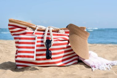 Stylish striped bag with visor cap, sunglasses and blanket on sandy beach near sea