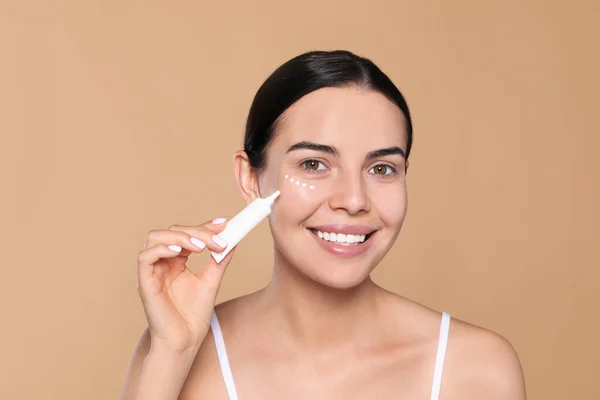 Beautiful Young Woman Applying Cream Skin Eye Beige Background — Zdjęcie stockowe
