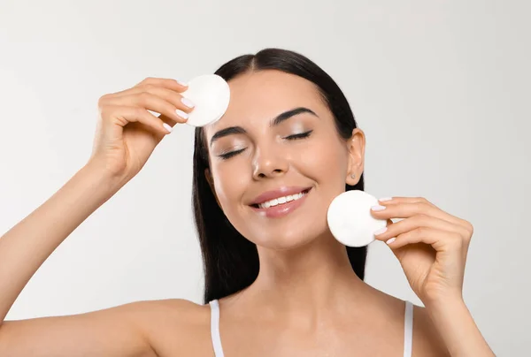 Young woman using cotton pads with micellar water on light grey background