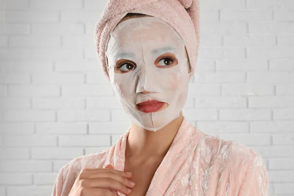 Young woman in bathrobe with cotton facial mask near white brick wall