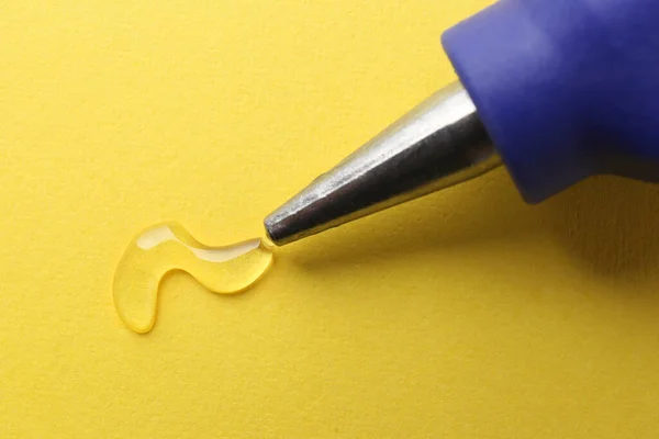 Melted glue dripping out of hot gun nozzle on yellow background, closeup