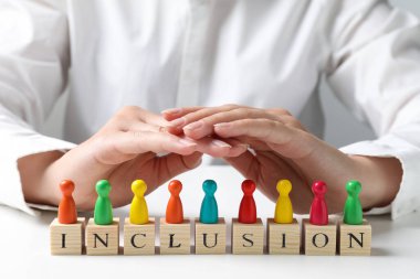 Woman protecting colorful pawns and wooden cubes with word Inclusion at white table, closeup