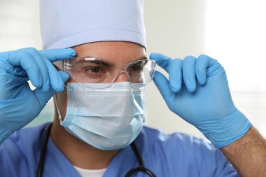 Koruyucu maskeli doktor, gözlüklü ve eldivenli, arka planda ışığa karşı.
