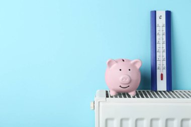 Piggy bank and thermometer on heating radiator against light blue background, space for text