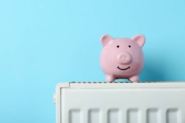 Piggy bank on heating radiator against light blue background, space for text