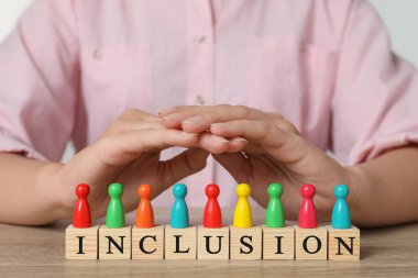 Woman protecting colorful pawns and wooden cubes with word Inclusion at table, closeup