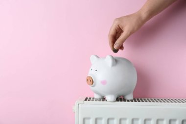 Woman putting coin into piggy bank on heating radiator against pink background, closeup. Space for text