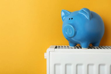 Piggy bank on heating radiator against orange background, space for text