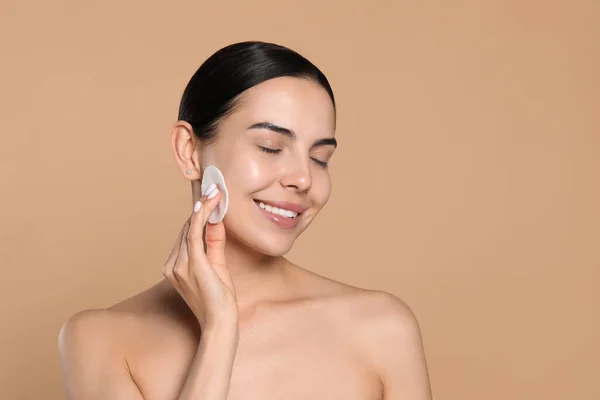 Young woman using cotton pad with micellar water on beige background, space for text