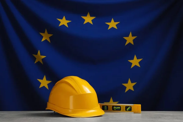 Yellow hard hat and building level on light grey table against flag of European Union