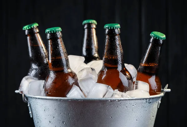 Metal bucket with bottles of beer and ice cubes on dark wooden background, closeup