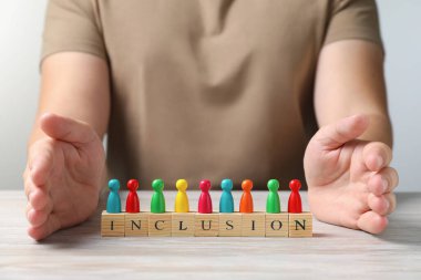 Woman protecting colorful pawns and cubes with word Inclusion at wooden table, closeup