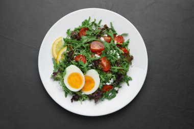 Delicious salad with boiled egg, arugula and tomatoes on black table, top view
