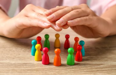 Woman sheltering colorful pawns at wooden table, closeup. Social inclusion concept