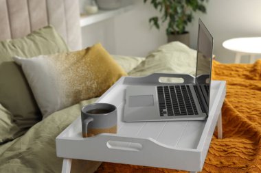 Modern dizüstü bilgisayarlı beyaz tepsi ve yatağın içinde bir fincan aromatik çay.