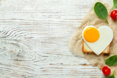 Heart shaped fried egg with toast, spinach and tomatoes on white wooden table, flat lay. Space for text