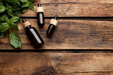 Bottles of basil essential oil with fresh leaves on wooden table, flat lay. Space for text