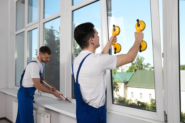 Workers with tube of sealant and suction lifters installing plastic windows indoors