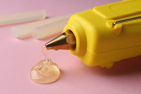 Melted glue dripping out of hot gun nozzle near sticks on pink background, closeup