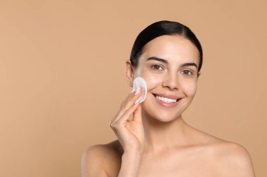 Young woman using cotton pad with micellar water on beige background, space for text