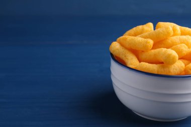 Bowl of tasty cheesy corn puffs on blue wooden table, space for text