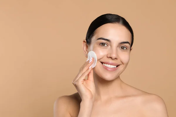 Young woman using cotton pad with micellar water on beige background, space for text