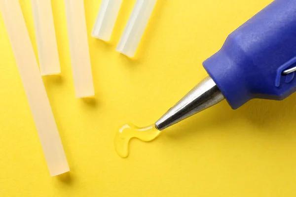 Melted glue dripping out of hot gun nozzle near sticks on yellow background, closeup