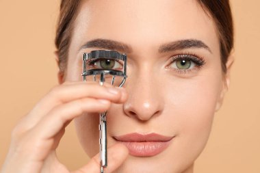 Woman using eyelash curler on beige background, closeup