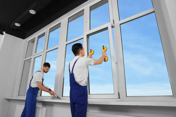 Workers with tube of sealant and suction lifters installing plastic windows indoors