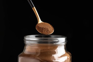 Spoon of instant coffee over jar against black background, closeup