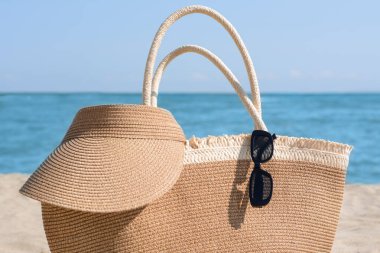 Stylish straw bag with visor cap and sunglasses on beach near sea, closeup
