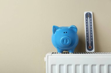 Piggy bank and thermometer on heating radiator against beige background, space for text
