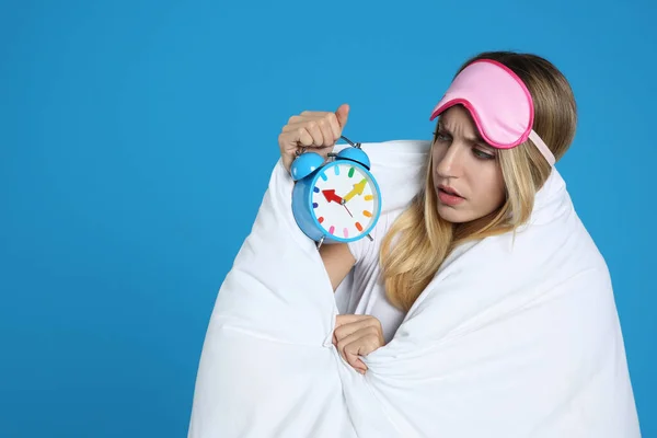 Emotional young woman in sleeping mask wrapped with blanket holding alarm clock on blue background