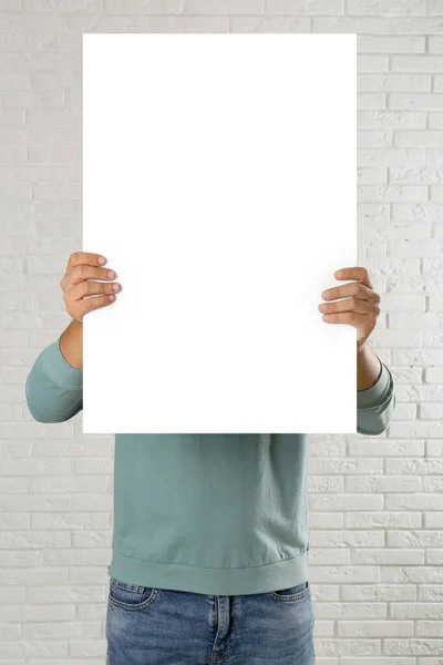 Man holding blank poster near white brick wall. Mockup for design