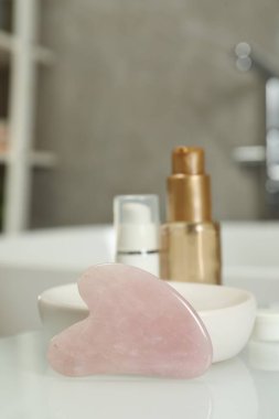 Rose quartz gua sha tool and cosmetic products on white table in bathroom