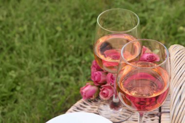 Picnic basket with glasses of delicious rose wine and flowers outdoors, space for text