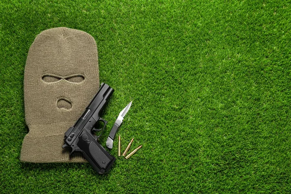 Flat lay composition with balaclava and weapons on green grass. Space for text