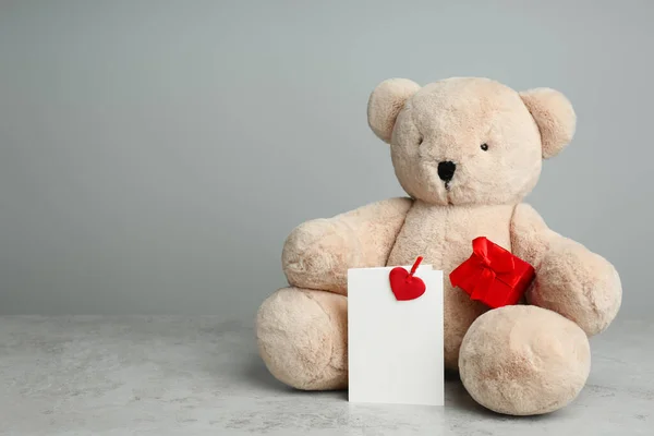 Cute teddy bear with gift box and blank card on light grey table, space for text. Valentine\'s day celebration