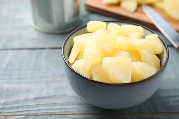 Pieces of delicious canned pineapple in bowl on light blue wooden table, closeup