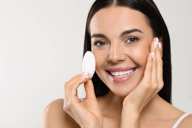 Young woman using cotton pad with micellar water on light grey background