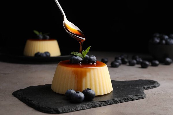 Pouring caramel onto delicious pudding with blueberries and mint on grey table against black background